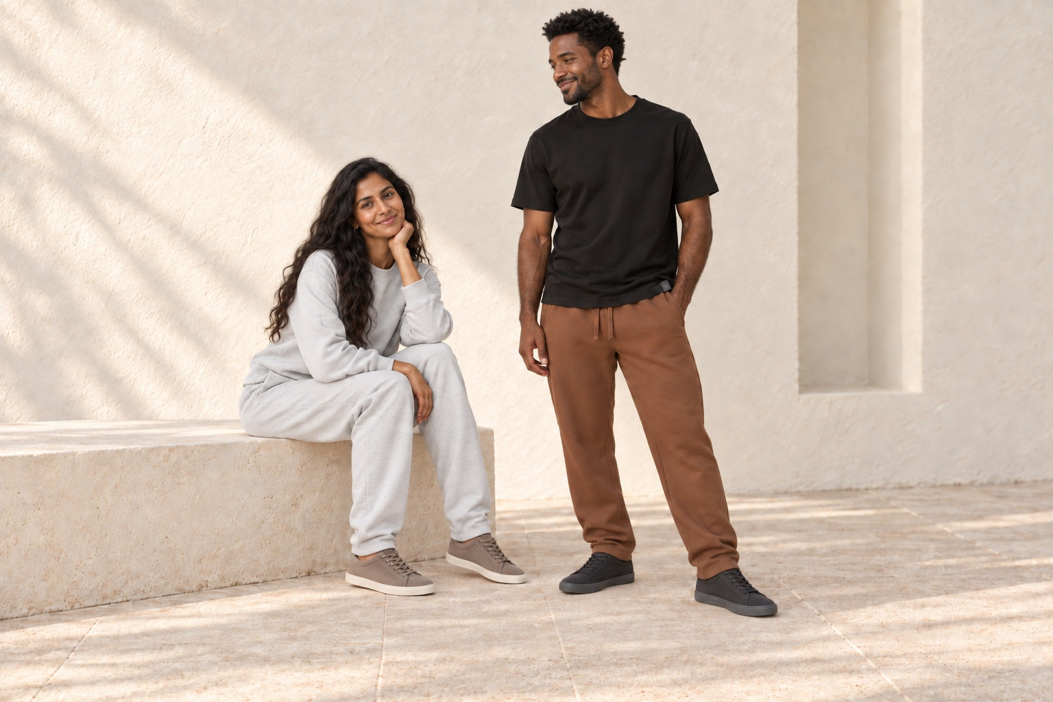 Two fashion models wearing a gray sweat set and a black T-shirt with brown sweatpants in a sunlit stone setting.